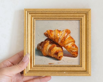 Könnte beinhalten: Ein kleines, gerahmtes Gemälde mit zwei goldbraunen Croissants. Das Gemälde hat einen goldenen Rahmen mit detailliertem Rand. Die Croissants sind mit detaillierten Pinselstrichen dargestellt, die ihre flockige Textur und satte Farbe zeigen. Der Hintergrund ist hellgrau.