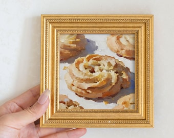 May include: A small, square, gold-framed painting featuring a close-up of several light brown, swirled pastries. The painting has a white background and is held by a person's hand.