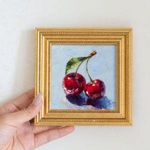 May include: A framed oil painting of two ripe cherries with a green leaf. The cherries are a deep red colour, and the background is a light blue. The frame is gold.