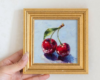 May include: A framed oil painting of two ripe cherries with a green leaf. The cherries are a deep red color, and the background is a light blue. The frame is gold.