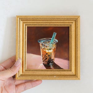 以下が含まれることがあります： バブルティーの絵画。透明なプラスチックカップに入った飲み物で、青いストローが付いています。バブルティーには、濃い茶色のタピオカパールと薄茶色の液体が入っています。フレームは金色です。