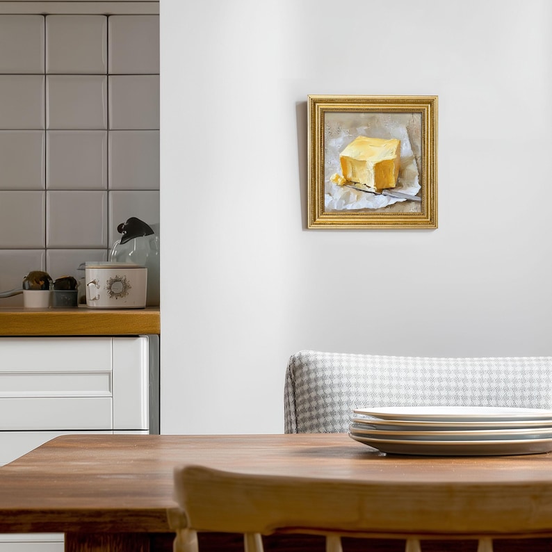 May include: A framed painting of a square block of yellow butter on parchment paper with a silver knife. The painting has a gold frame and is hanging on a white wall. The painting is in a dining room setting.