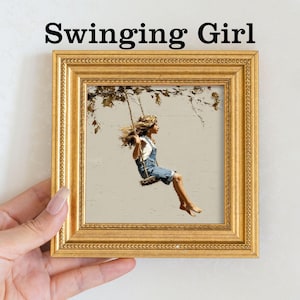 Pu&ograve; includere: Un'opera d'arte incorniciata intitolata "Swinging Girl" raffigura una giovane persona su un'altalena. L'opera d'arte &egrave; in una cornice dorata. La persona indossa una tuta di jeans e una camicia bianca. Lo sfondo &egrave; di colore neutro con rami di alberi.