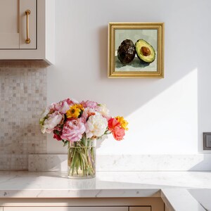 Op de afbeelding: Een ingelijst schilderij van twee avocado's, &eacute;&eacute;n heel en &eacute;&eacute;n gehalveerd, met een gouden lijst. Een boeket roze, gele en oranje bloemen in een heldere glazen vaas staat op een wit aanrecht. De keuken heeft een neutraal kleurenpalet.