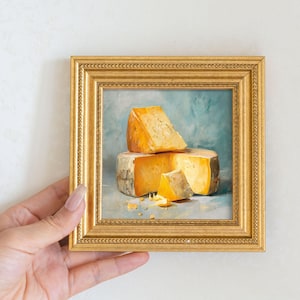 Peut inclure: Peinture encadr&eacute;e en or repr&eacute;sentant une nature morte de fromage. Le fromage est d'un orange vif, avec une part coup&eacute;e. Le cadre dor&eacute; a une bordure d&eacute;taill&eacute;e. L'arri&egrave;re-plan est bleu et vert doux. Une main tient le cadre.