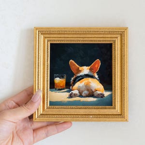 K&ouml;nnte beinhalten: Gerahmtes Gem&auml;lde eines Corgi-Hundes, der von hinten gesehen liegt, mit einem Glas bernsteinfarbener Fl&uuml;ssigkeit. Das Gem&auml;lde befindet sich in einem goldenen Rahmen mit detailliertem Rand. Der Corgi hat orange-wei&szlig;es Fell.