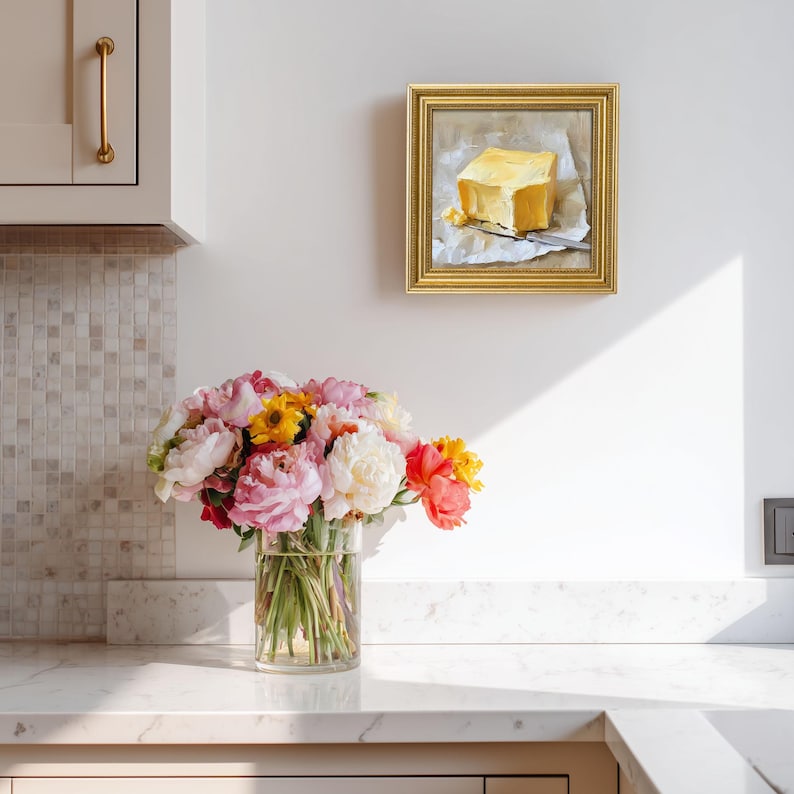 May include: A framed oil painting of a yellow butter block on parchment paper with a knife. A clear glass vase holds a bouquet of pink, yellow, and red flowers. The painting hangs on a white wall above a marble countertop.
