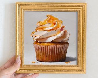 May include: A framed painting of a cupcake with white frosting and orange caramel drizzle. The cupcake sits in a brown paper liner. The painting is in a gold frame.