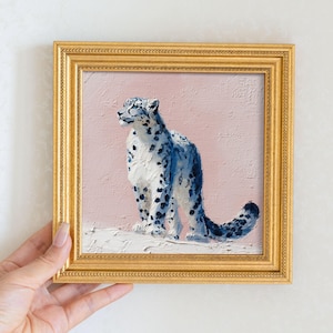 May include: Square framed painting of a snow leopard. The artwork features a blue and white snow leopard against a pink background. The frame is gold. The painting is held by a hand.