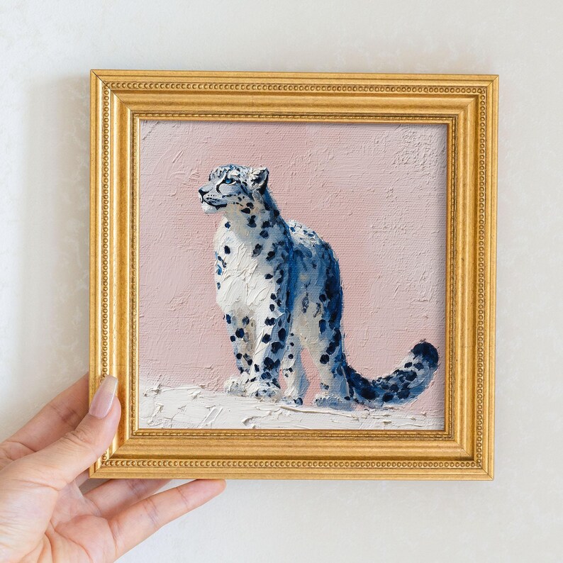May include: Square framed painting of a snow leopard. The artwork features a blue and white snow leopard against a pink background. The frame is gold. The painting is held by a hand.