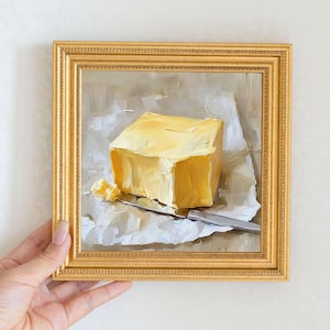 May include: A framed oil painting of a yellow butter block on white paper with a silver knife. The painting has a gold frame and is held by a hand. The artwork is a still life.