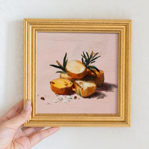 May include: An oil painting in a square gold frame depicting several halved yellow potatoes with green sprigs of rosemary. The artwork features visible brushstrokes and a textured application of paint against a solid light pink background.