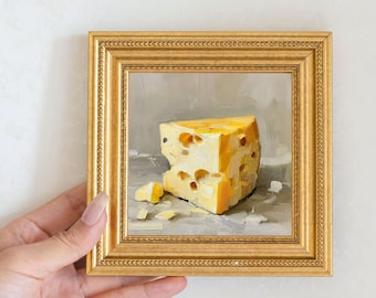 May include: A framed painting of a wedge of yellow cheese with holes. The painting is in a gold frame and is held by a hand. The cheese is on a gray surface.