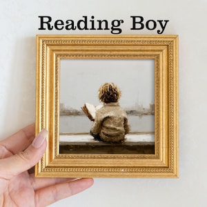 Pu&ograve; includere: Una stampa incorniciata intitolata "Reading Boy" raffigura un bambino intento a leggere un libro, seduto vicino a una finestra con vista su un paesaggio urbano. L'opera &egrave; racchiusa in una cornice dorata e ornata, con il testo "Reading Boy" in alto.