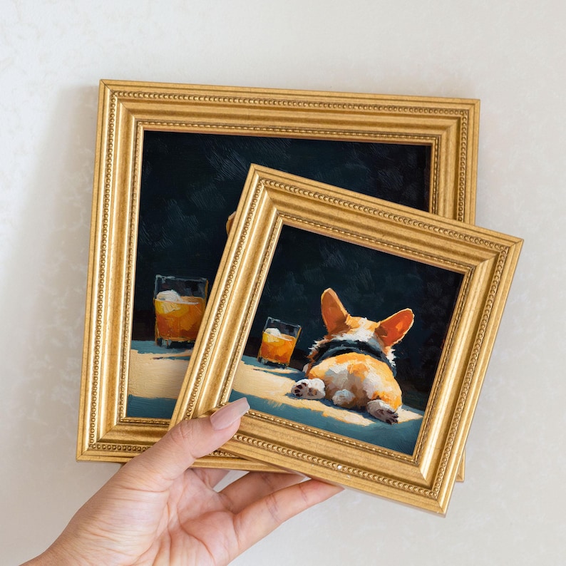 K&ouml;nnte beinhalten: Zwei gerahmte Kunstdrucke mit goldfarbenen Rahmen. Das Kunstwerk zeigt einen Corgi-Hund und ein Glas bernsteinfarbener Fl&uuml;ssigkeit. Der Corgi liegt, und der Hintergrund ist dunkel. Die Rahmen haben verzierte Details.