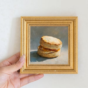 May include: A small, square, gold-framed painting depicts a biscuit sandwich with a golden-brown exterior and a filling of orange preserves. The painting is held by a hand with long nails. The background is a soft, muted gray.