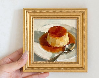May include: A framed painting of a flan dessert on a white plate with a silver spoon. The flan is topped with a caramel sauce. The frame is gold with a detailed border.