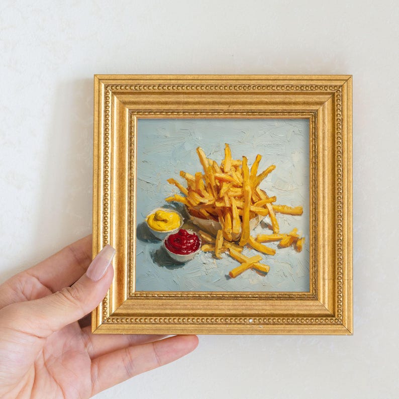 May include: A framed painting of golden chips with small bowls of ketchup and mustard. The painting has a light blue background and is in a gold frame. The painting is held by a person.