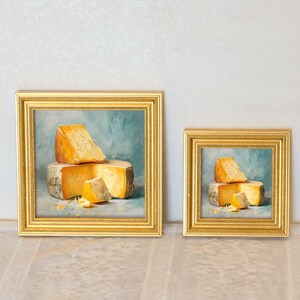 Peut inclure: Deux tableaux encadr&eacute;s avec des cadres dor&eacute;s. Chaque peinture repr&eacute;sente une nature morte de morceaux de fromage sur fond bleu. Le fromage est un m&eacute;lange de teintes jaunes et orang&eacute;es. Les peintures sont expos&eacute;es sur une surface blanche.