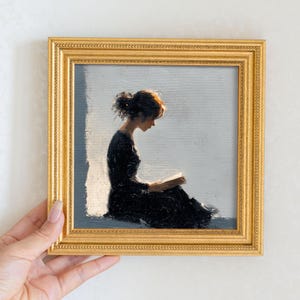 May include: A framed painting depicts a person in a black dress absorbed in reading a book. The artwork is set against a light gray background with a golden frame. The person is seated, illuminated by a soft light.