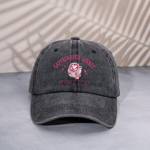 Puede incluir: Gorra de béisbol gris con la inscripción "Caffeinated Disney Moms Club" bordada en rosa. El diseño incluye una taza de café con un corazón y estrellas. La gorra tiene una visera curva y un aspecto desgastado.