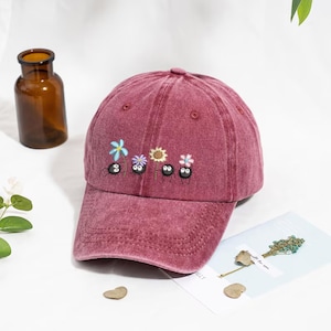 Könnte beinhalten: Burgunderfarbene Baseballkappe mit gebogenem Schirm. Die Kappe hat ein gesticktes Design aus Blumen und kleinen, schwarzen, runden Kreaturen. Die Kappe besteht aus einem weichen, strukturierten Stoff und hat ein klassisches Sechs-Panel-Design.