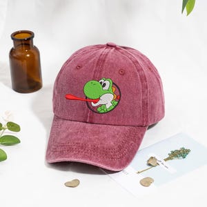 Peut inclure: Casquette de baseball marron avec une image brodée d'un personnage Yoshi vert avec une langue rouge. La casquette a une visière incurvée et un design classique à six panneaux. Fabriquée en tissu doux et lavé.
