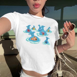 Può includere: T-shirt bianco girocollo con un design a cartoni animati di pesci in sacchetti blu che galleggiano nell'acqua. La grafica include una stella marina, un pesce pagliaccio e altre creature marine. La maglietta è realizzata in materiale morbido e leggero.