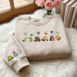 Op de afbeelding: Beige sweatshirt met geborduurde cartoon dieren met hartvormige ballonnen. De mouw heeft extra geborduurde ontwerpen. De sweatshirt wordt weergegeven op een pluizig wit oppervlak, met een raam en bloemen op de achtergrond.