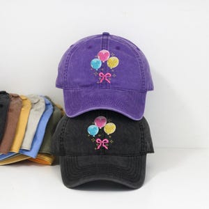 Op de afbeelding: Twee baseballpetten, één paars en één zwart, zijn op elkaar gestapeld. Elke pet heeft een geborduurd ontwerp van drie ballonnen in roze, blauw en geel, met een roze strik. De petten zijn gemaakt van een zachte, gewassen stof.