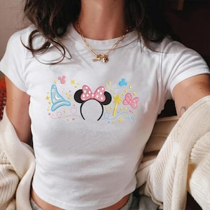 Puede incluir: Camiseta blanca con un diseño bordado colorido que presenta una diadema de orejas de Minnie Mouse con un lazo rosa, un sombrero de mago, una varita mágica y otros elementos caprichosos. La camiseta es de manga corta.