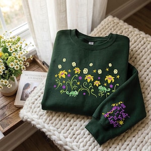 May include: A dark green crewneck sweatshirt featuring a graphic print of yellow flowers, green chameleons, and floating lanterns. The sleeve displays the purple and yellow text, And at last I see the light, accompanied by a small sun and lantern icon.