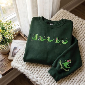 Può includere: Felpa verde scuro con lucertole verdi ricamate sul petto e sulla manica. La manica presenta la scritta "lizard lizard lizard". La felpa è mostrata su una superficie a maglia color crema.