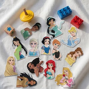 Könnte beinhalten: Eine Sammlung von dreizehn gestanzten Aufklebern mit verschiedenen animierten Prinzessinnen und Heldinnen, darunter Figuren mit langem blondem, braunem, schwarzem und rotem Haar, sowie ein gelber Kreisel, ein blauer und ein roter Baustein.