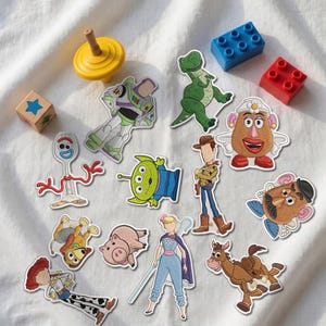 Könnte beinhalten: Eine Sammlung von gestickten Aufnähern mit Charakteren aus Toy Story, darunter Buzz Lightyear, Rex, Forky, ein Alien, Woody, Bo Peep, Bullseye, Slinky Dog, Hamm sowie Mr. und Mrs. Potato Head, ergänzt durch einen gelben Kreisel und zwei Plastikbausteine.
