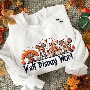 May include: A white crewneck sweatshirt featuring an embroidered design of a castle, a water tower, and a geodesic sphere with Mickey Mouse ears. The text Walt Disney World is embroidered in black below the icons, accented by orange autumn leaves.