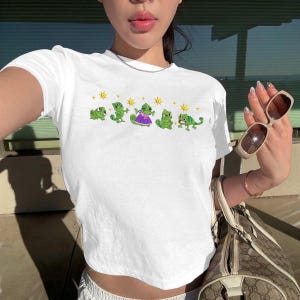 Könnte beinhalten: Weißes Crop-Top mit einer Reihe von grünen Cartoon-Chamäleons und gelben Sternen. Ein Chamäleon trägt ein lila Kleid. Das Oberteil ist ein lässiger Stil mit kurzen Ärmeln.