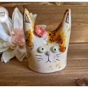 May include: A ceramic cat figurine with a cream-coloured base, orange and brown patches, and a small pink flower on its head. The cat has two pointed ears, wide green eyes, a small pink nose, and etched whiskers. The text PARRYS POTTERY is visible.