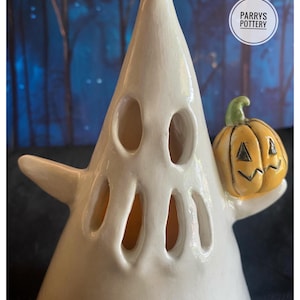 May include: A white ceramic ghost figurine with cut-out eyes and a mouth, holding a small yellow jack-o'-lantern with a green stem. The ghost has a conical shape and a smooth, glossy finish. The text PARRYS POTTERY is visible in a white circle.