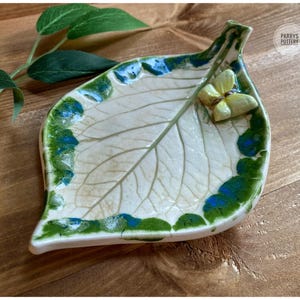 May include: A ceramic leaf-shaped dish with a cream-coloured centre featuring detailed vein textures. The edges are decorated with green and blue glaze, and a small, pale yellow ceramic butterfly is attached to the surface. Text reads PARRYS POTTERY.