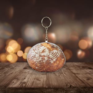 Op de afbeelding: Een sleutelhanger in de vorm van een rond brood, met een zilveren sleutelring en ketting. De sleutelhanger heeft een gedetailleerde afbeelding van een brood met een knapperige, bruine buitenkant en een witte binnenkant. De achtergrond is wazig met warme, gouden bokeh-lichten.