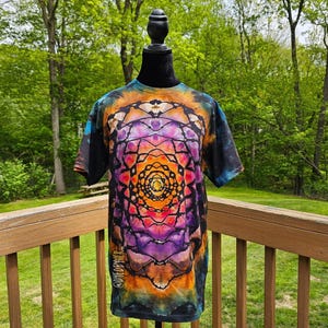 May include: A short-sleeved, tie-dye t-shirt with a vibrant, symmetrical design in shades of orange, purple, and blue. The shirt is displayed on a black mannequin against a backdrop of green trees. The text "@DYEDBYHER" is visible on the lower left side.