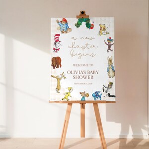 May include: A baby shower welcome sign on a wooden easel. The sign features a cream-colored background with a light plaid pattern and illustrations of storybook characters. Text reads "a new chapter begins" and "Olivia's Baby Shower, September 21, 2026."