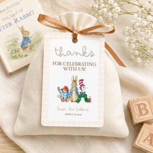 May include: A cream-colored fabric drawstring pouch with a brown ribbon tied at the top. A rectangular tag features the text "thanks FOR CELEBRATING WITH US!", illustrations of Paddington Bear, Peter Rabbit, and The Very Hungry Caterpillar, "love, the bakers", and "MARCH 14, 2026".