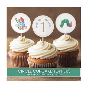 Puede incluir: Tres cupcakes con glaseado blanco están cubiertos con adornos circulares. Uno presenta a Paddington Bear, otro tiene el número 1 y el texto "Happy Birthday Anna", y el tercero muestra una oruga verde. El texto "CIRCLE CUPCAKE TOPPERS STORYBOOK BIRTHDAY DECOR" está en la parte inferior.