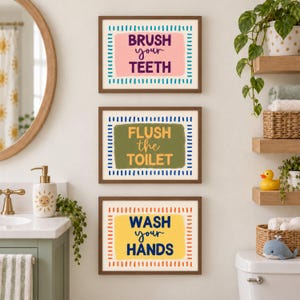 May include: Three rectangular wall art prints in brown frames. The top print has a pink background with blue dashes and the text BRUSH your TEETH. The middle print has a green background with blue dashes and the text FLUSH the TOILET. The bottom print has a yellow background with orange dashes and the text WASH your HANDS.