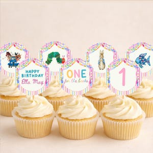 May include: A set of round cupcake toppers featuring colorful illustrations of storybook characters, including a blue truck with animals, a green caterpillar, a rabbit, and a blue fish, with text reading Happy Birthday Ella May and ONE for the books.