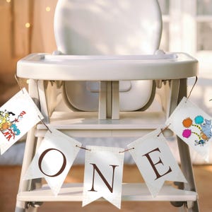 May include: A white high chair with a banner that spells "ONE". The banner has brown letters and is decorated with illustrations of characters from Dr. Seuss books. The high chair is set up for a first birthday celebration.
