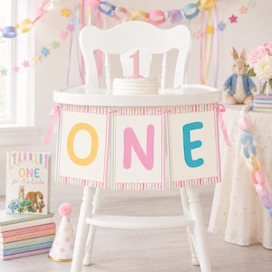 May include: A white high chair decorated with a banner featuring the word ONE in yellow, pink, and blue letters. A small white cake with a pink glittery number one topper sits on the tray. A book titled ONE for the books sits nearby.