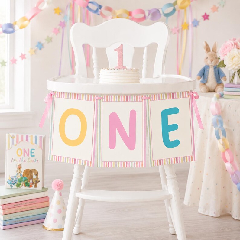 May include: A white high chair decorated with a banner featuring the word ONE in yellow, pink, and blue letters. A small white cake with a pink glittery number one topper sits on the tray. A book titled ONE for the books sits nearby.